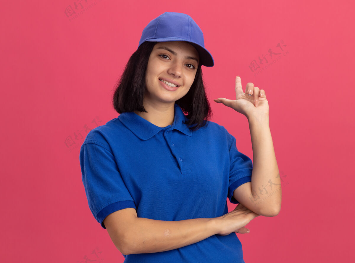 制服身着蓝色制服 头戴鸭舌帽的快乐的年轻送货女孩微笑着指着粉红色的墙上站着的指数标杆帽子送货员目录