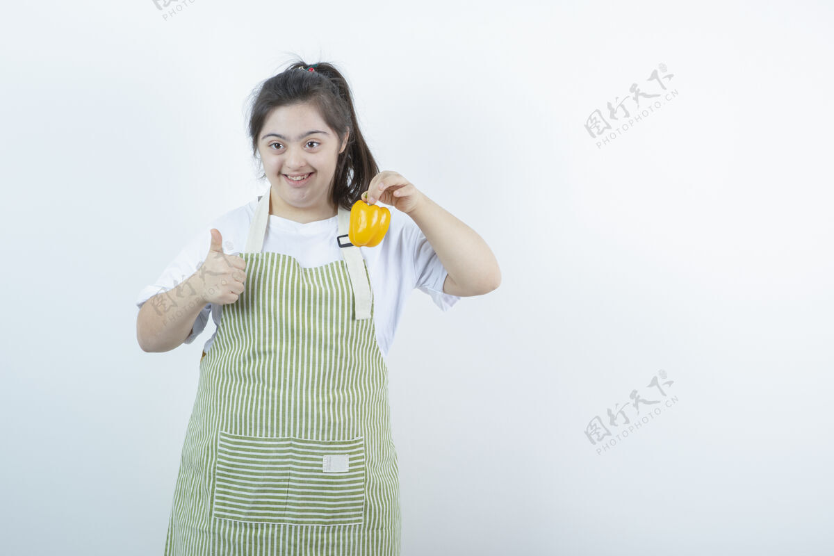 食物穿着格子围裙的年轻漂亮女孩 手里拿着黄色的甜椒 竖起大拇指残疾成人年轻人