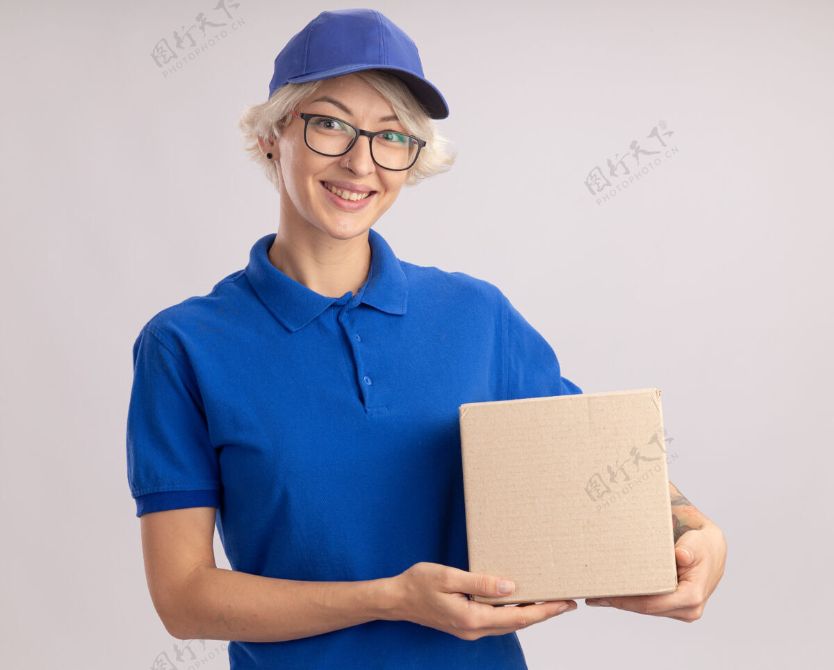 欢呼身着蓝色制服 戴着眼镜 拿着纸板箱 站在白墙上开心地微笑着的年轻送货员递送站着戴着
