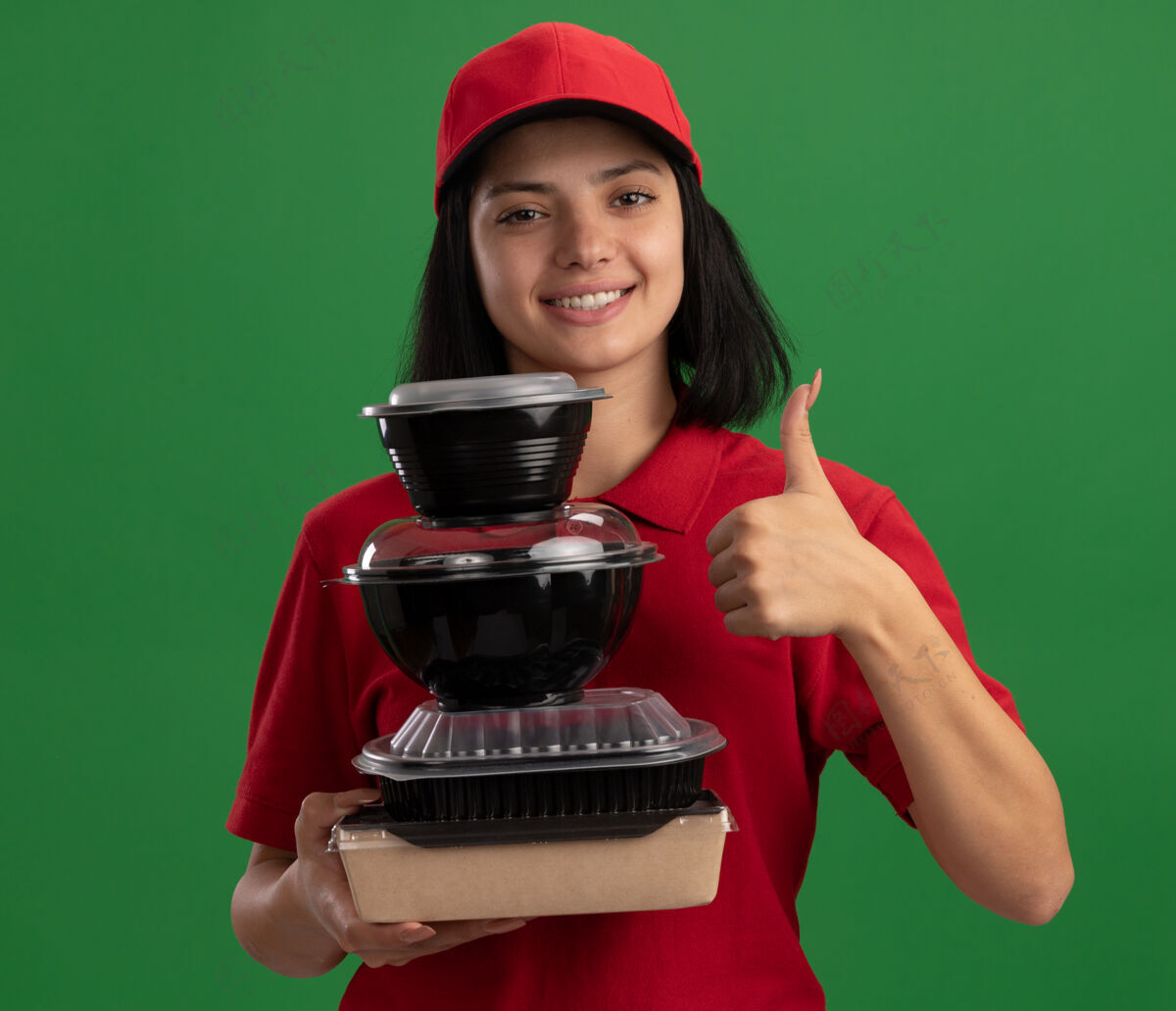 站着穿着红色制服 戴着帽子 手里拿着一叠食品包装 微笑着 友好地竖起大拇指 站在绿色的墙上 快乐的年轻送货女孩帽子微笑制服