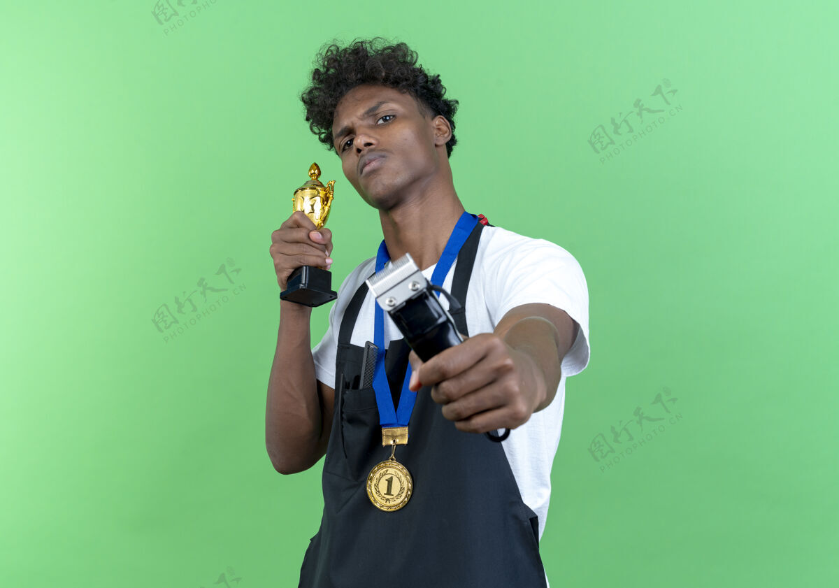 穿自信的年轻美国黑人男理发师穿着制服 拿着奖牌 拿着理发器对着镜头 优胜者杯被隔离在绿色背景上奖牌优胜者手持