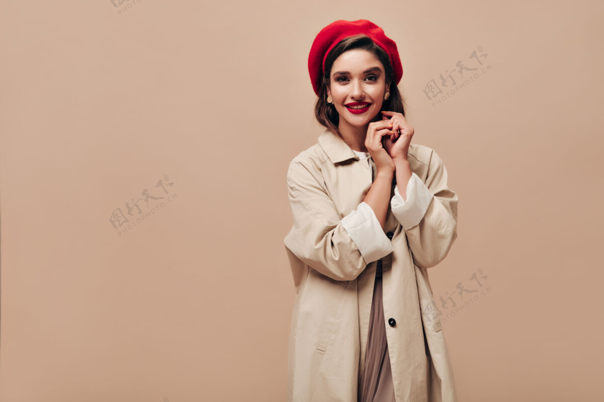 时尚心情好的女士看着米色背景的相机美丽的微笑着的女人 戴着红色贝雷帽 戴着耳环 穿着长外套 大而明亮的嘴唇女性巴黎优雅