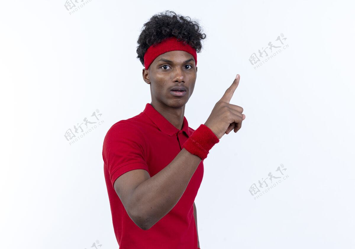 穿着令人印象深刻的年轻黑人美国运动男子戴头带和腕带点在一边孤立的白色背景与复制空间头带腕带尖