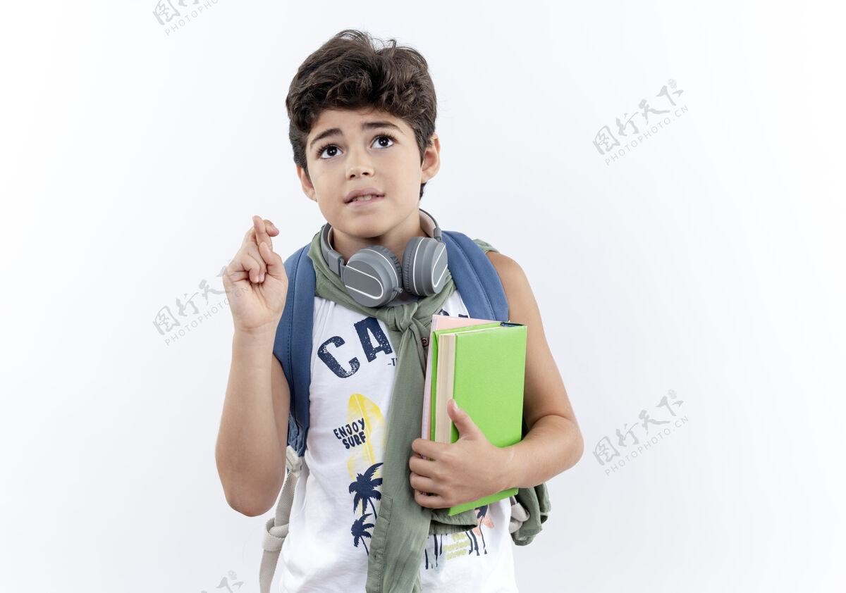 关心抬头望着那个戴着背包和耳机 拿着书 交叉着手指 孤立在白色背景上的小男孩小小学生抱