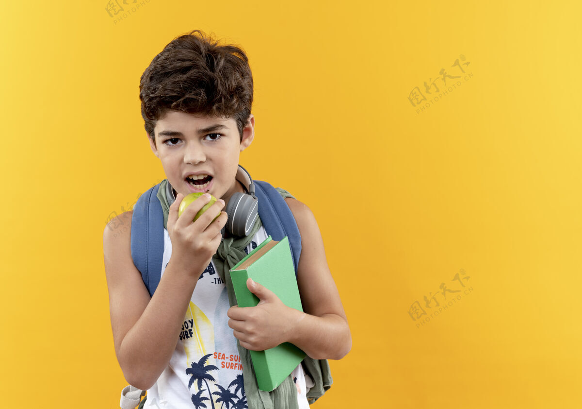 小看着镜头 一个背着书包 戴着耳机 拿着书 咬着黄色背景上孤立的苹果的小男孩耳机包咬