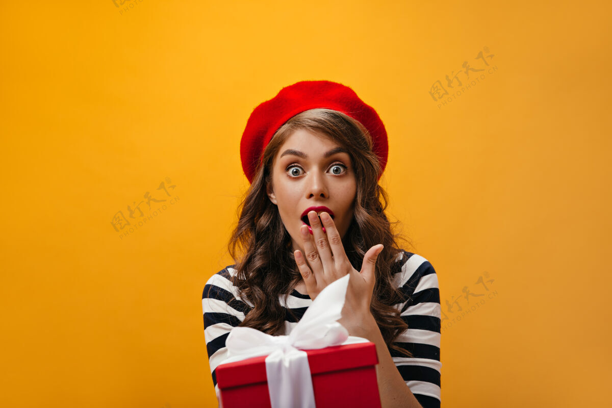 女士蓝眼睛的女人看起来很震惊 得到了礼物一个穿着红色贝雷帽和条纹衬衫的卷发女孩拿着礼物盒牛仔裙姿势休闲