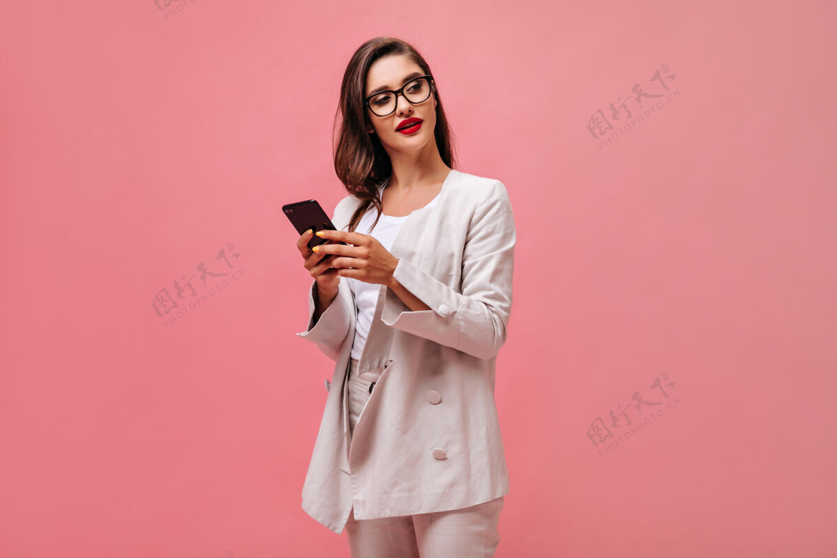 女孩穿着时髦服装 戴着眼镜的黑发女人拿着智能手机摆姿势红唇 白西装的商务小姐拿着手机时尚女士卷发