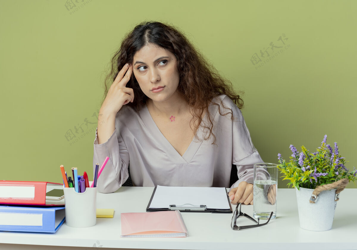 办公桌看着一边思考的年轻漂亮的女上班族坐在办公桌旁 把手指放在橄榄色背景下孤立的前额上橄榄思考工人