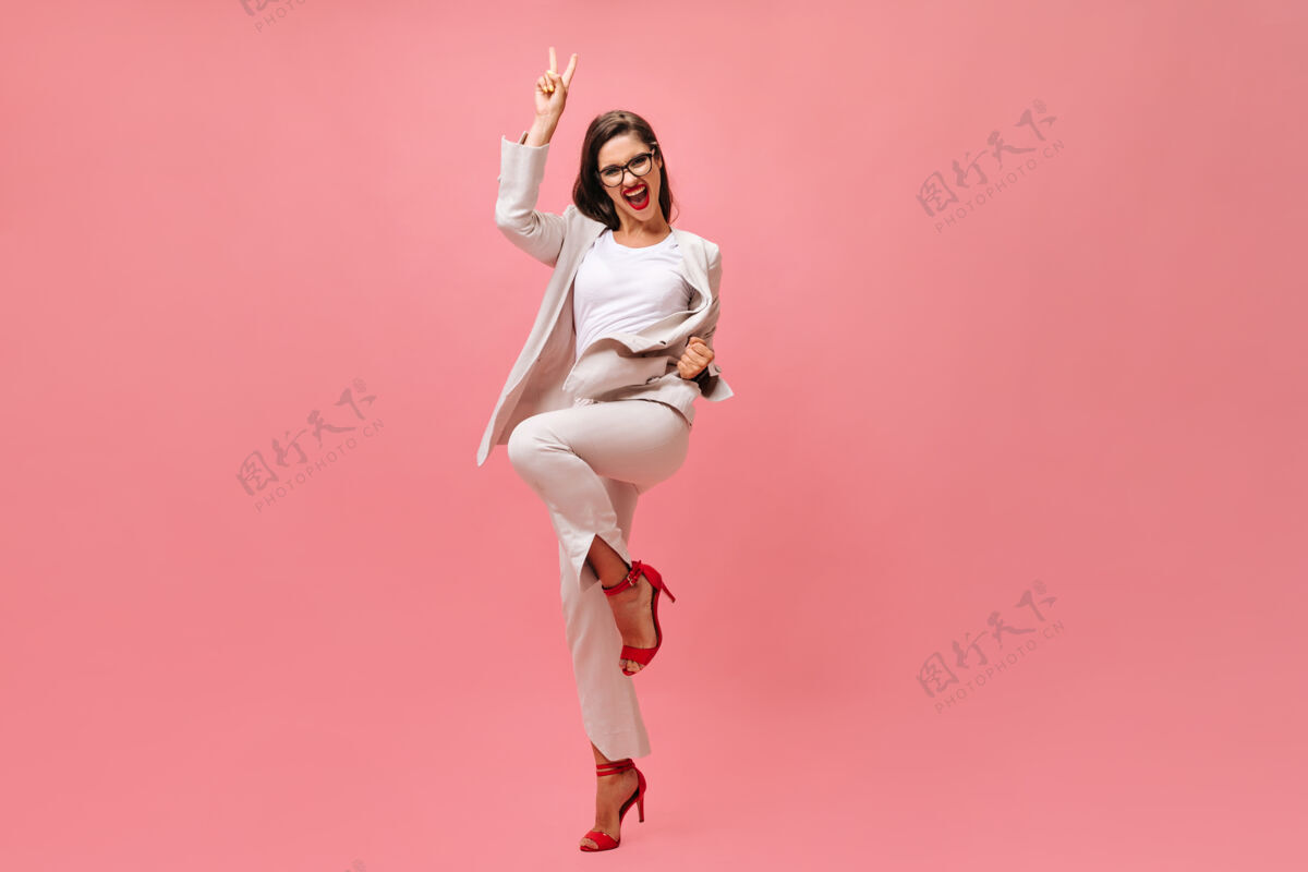 站立穿西装的快乐女人在粉色背景上显示和平标志穿着时髦服装的快乐美女在镜头前欢欣鼓舞平板女士姿势
