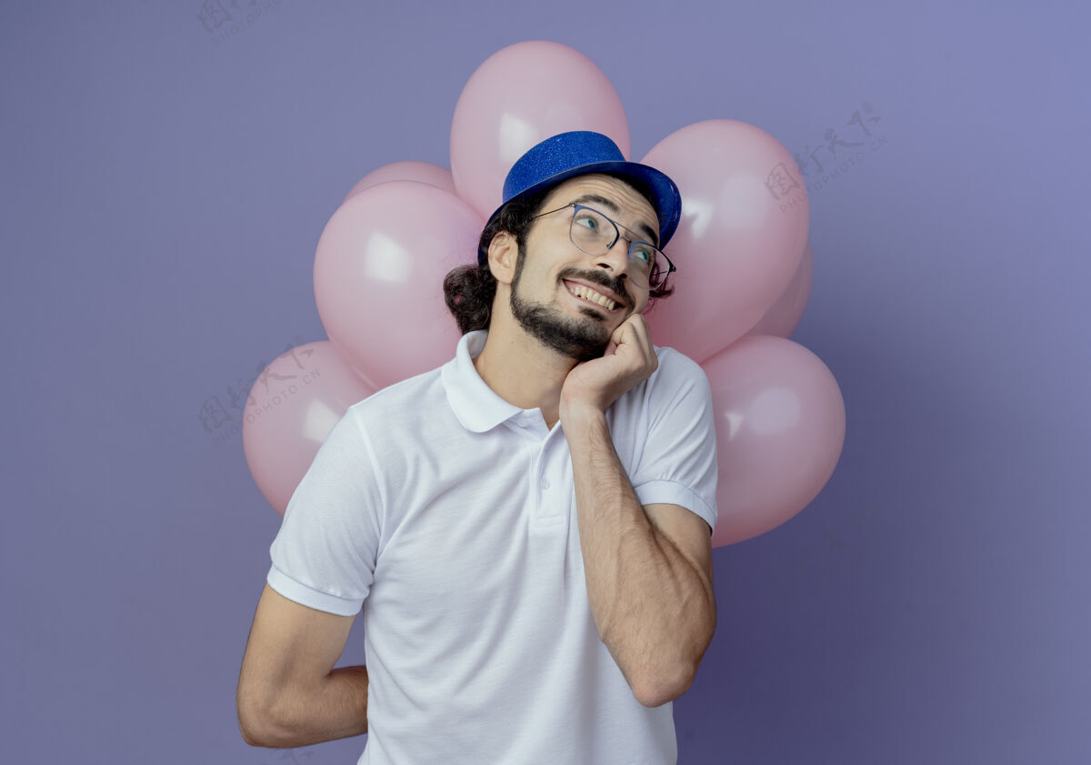 请穿看着身边高兴的帅哥戴着眼镜和蓝色帽子站在气球前 手放在下巴上孤立的紫色背景上壁板前面站