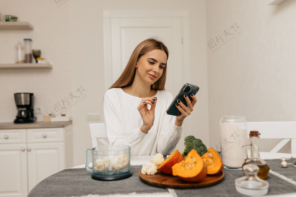 金发一位留着浅棕色头发的欧洲女士正在笑着 看着智能手机里的菜谱这位正在准备用南瓜和花椰菜做饭的年轻女士 爱好大笑人