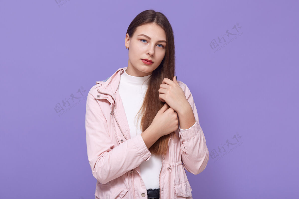 女性悲伤的年轻女子一边抚摸着头发 一边直视着 脸上露出不安的表情 穿着淡粉色夹克和白色高领毛衣 孤立地站在淡紫色的墙上心烦意乱卷发新鲜