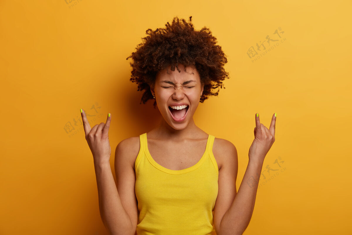 手势积极快乐的女人做重金属招牌 在音乐摇滚节上玩得开心 大声喊叫 闭上眼睛 积极地做手势 穿着休闲服 隔着黄色的墙壁摇滚宝贝非洲音乐摇滚