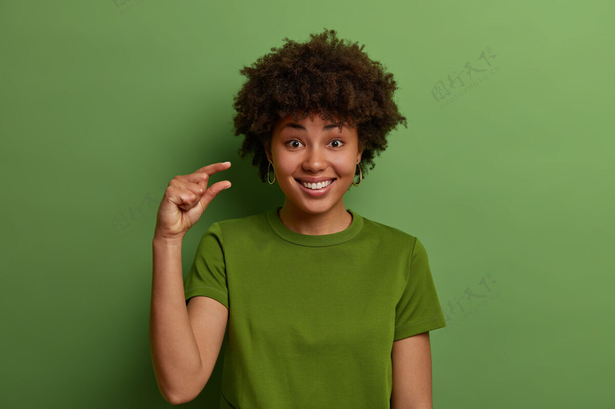 请积极开朗的黑皮肤卷发女人用手指塑造很少的东西 表现出小幅降价或降薪 手势不是大对象 咧嘴微笑 绿色占上风牙齿心情摄影