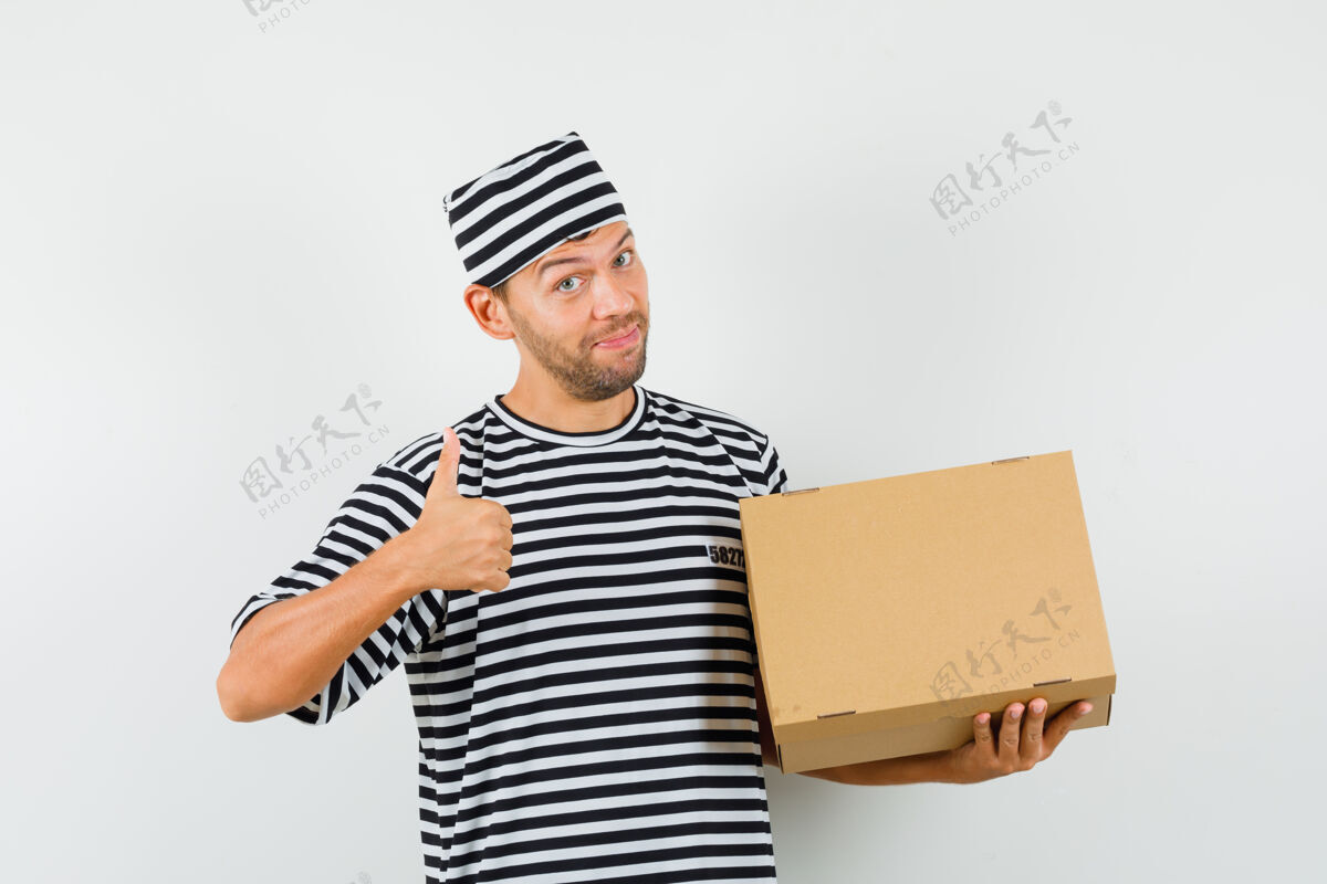 高兴一个戴着条纹t恤帽子的年轻人拿着一个纸板箱 竖起大拇指 看上去很高兴条纹休闲男性
