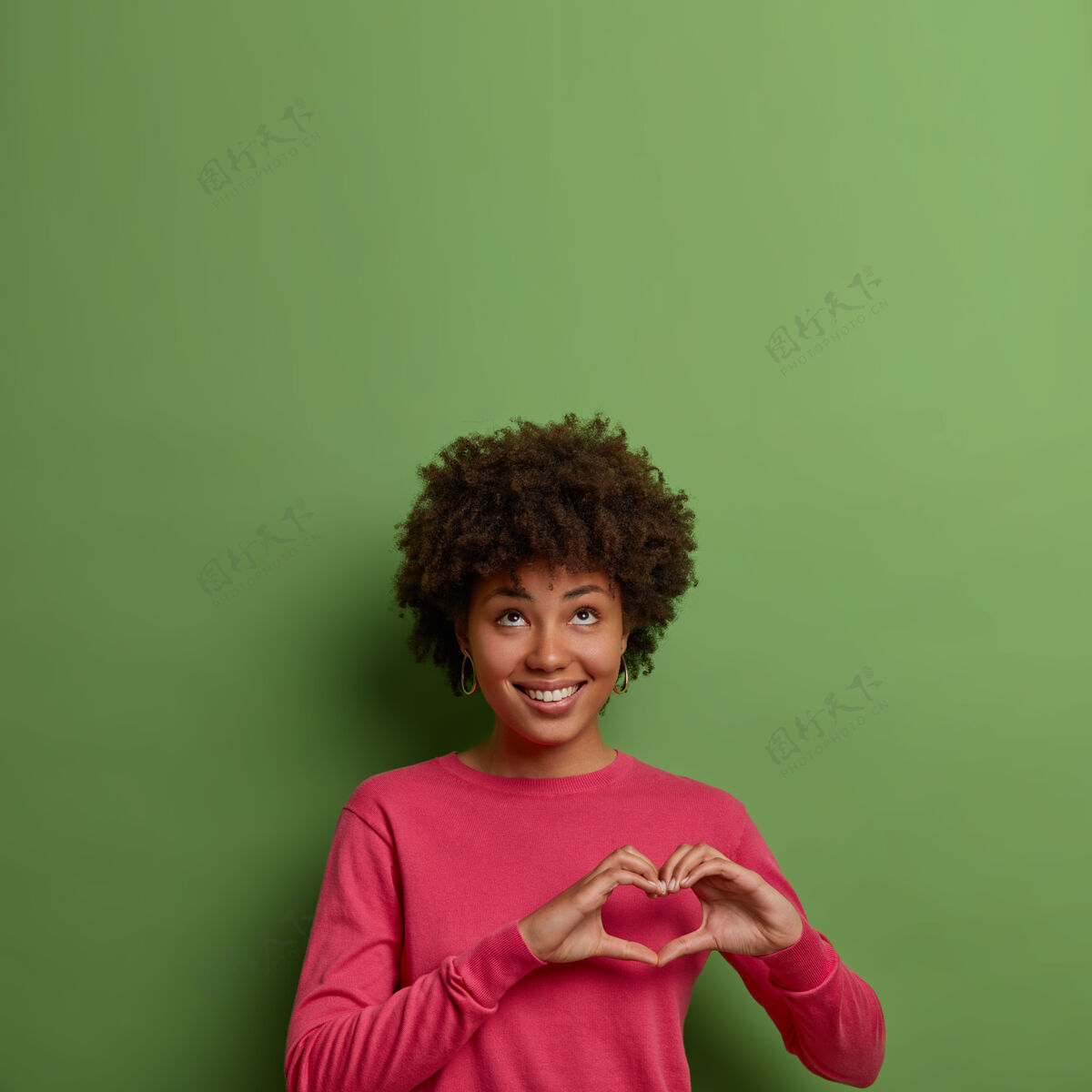 快乐一幅令人愉快的美籍黑人女性肖像 胸前有心形标志 表达爱意和关怀 抬头看去 身穿粉色套头衫 对着绿色墙壁摆姿势 向上腾空套头衫卷发显示