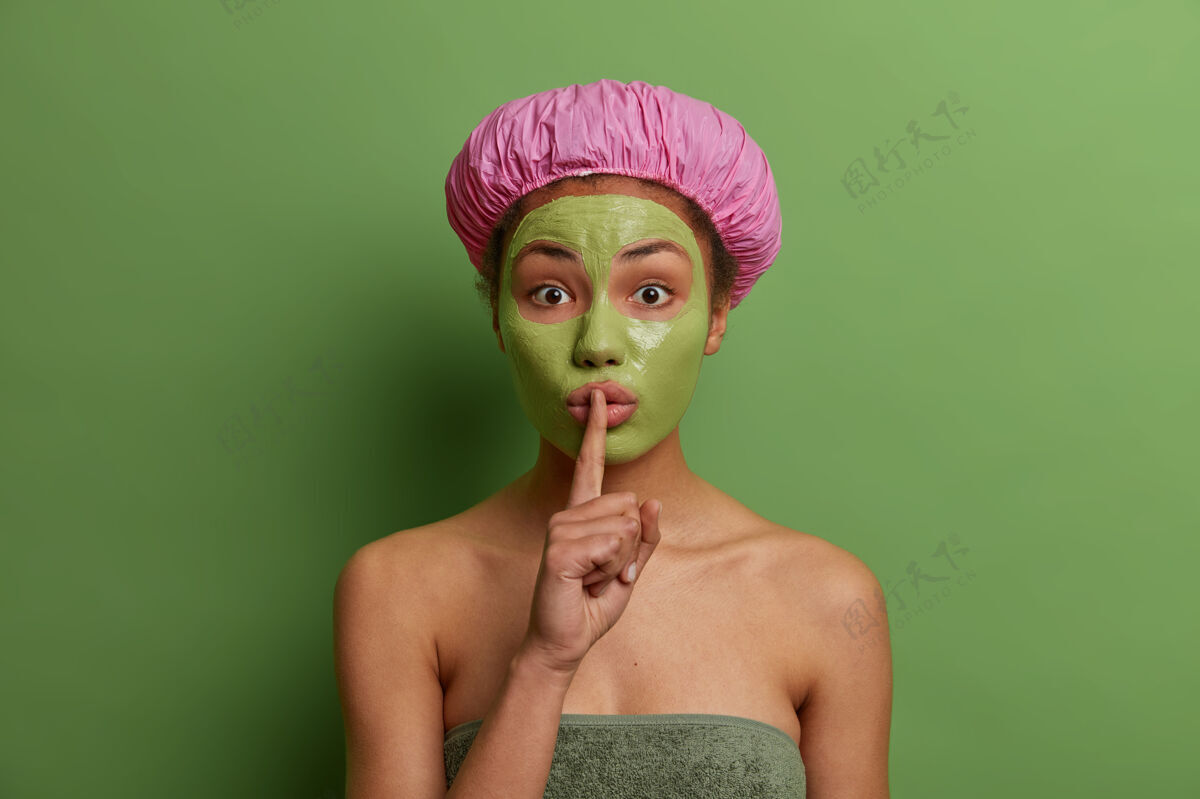 面部惊讶的年轻女模特 脸上戴着绿色的剥皮面膜 做着沉默的手势 讲述美容师的秘密 戴着浴帽 对着生动的墙壁摆姿势美容养颜理念泥淋浴美容师