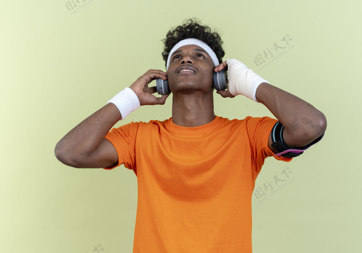 年轻看着年轻的美国黑人运动男子戴着头带和腕带和电话臂带耳机隔离在绿色的背景耳机腕带电话