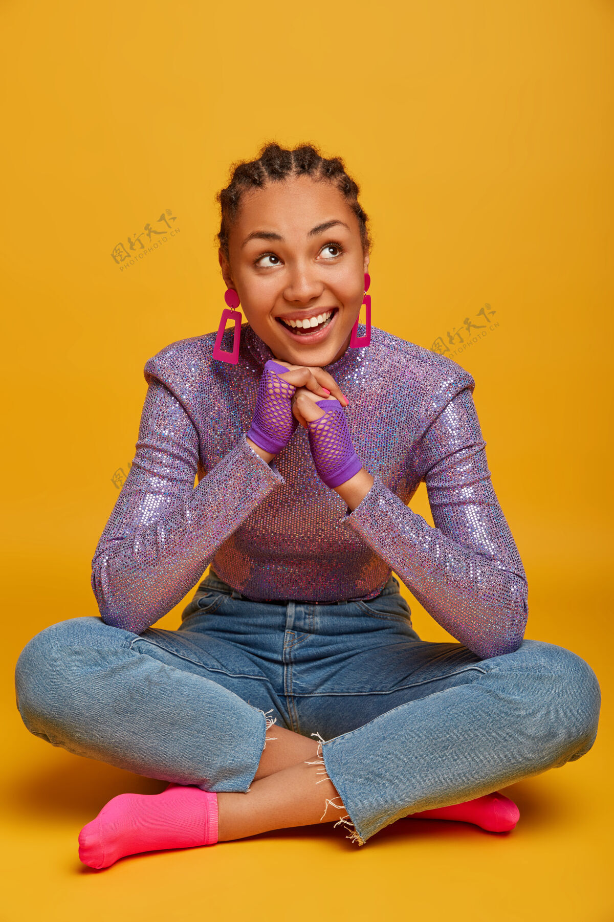 高兴快乐的美籍黑人年轻女子 双手放在下巴下 坐在黄色的墙上 穿着时髦的衣服 戴着运动手套 面带微笑请姿势幻想