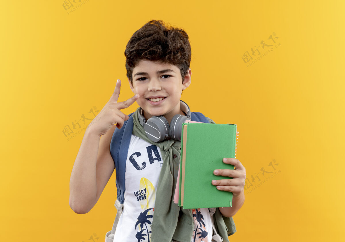 耳机高兴的小男生背着书包 戴着耳机 拿着书 在黄色背景上表现出与世隔绝的和平姿态和平男孩显示
