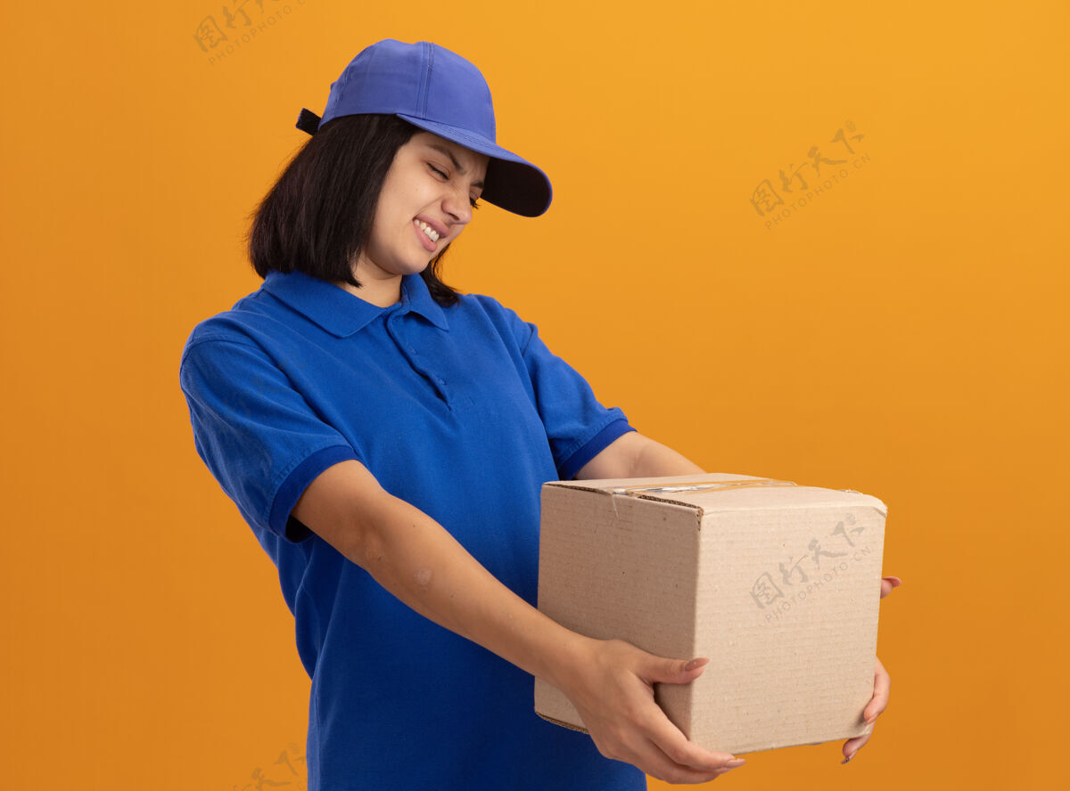 恶心身穿蓝色制服 戴着帽子 拿着纸板箱的年轻送货女孩站在橙色的墙上 带着厌恶的表情看着它女孩站着年轻