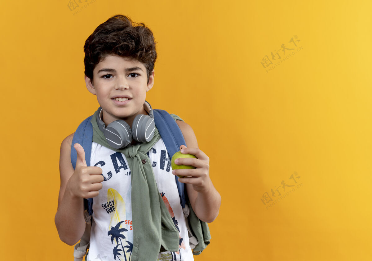 抱高兴的小男孩戴着背包和耳机拿着苹果他的大拇指孤立的黄色背景小请拇指