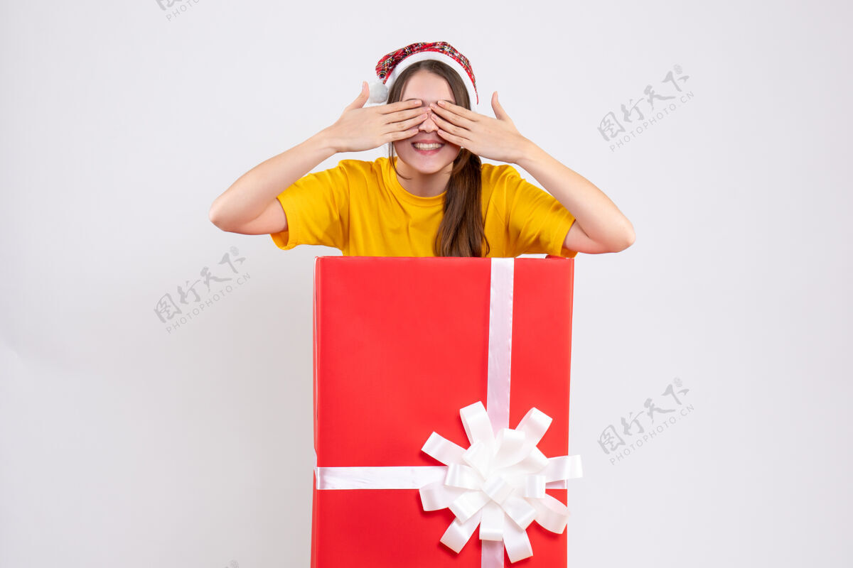 大正面图可爱的女孩戴着圣诞帽遮住眼睛 双手站在圣诞礼物后面证人圣诞老人购物
