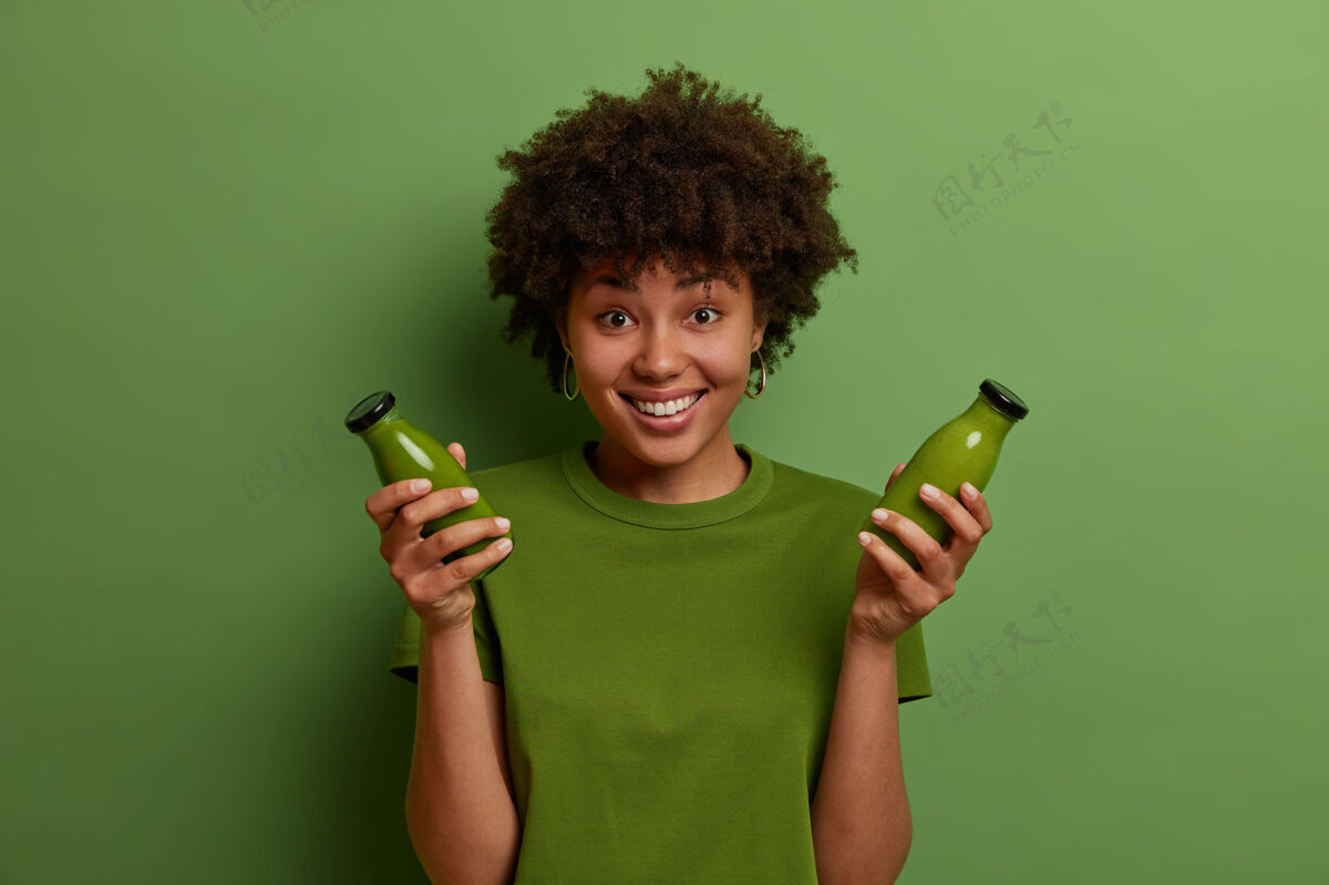 玻璃快乐的黑皮肤女孩拿着玻璃瓶和生的绿色排毒蔬菜冰沙 引导健康的生活方式 坚持素食 感觉清爽和高兴单色拍摄人民和健康享受心情冰沙