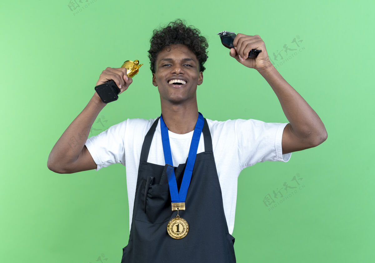 优胜者快乐的年轻黑人美国男理发师穿着制服和奖牌提高冠军杯与剪发隔离在绿色背景奖牌奖杯理发师