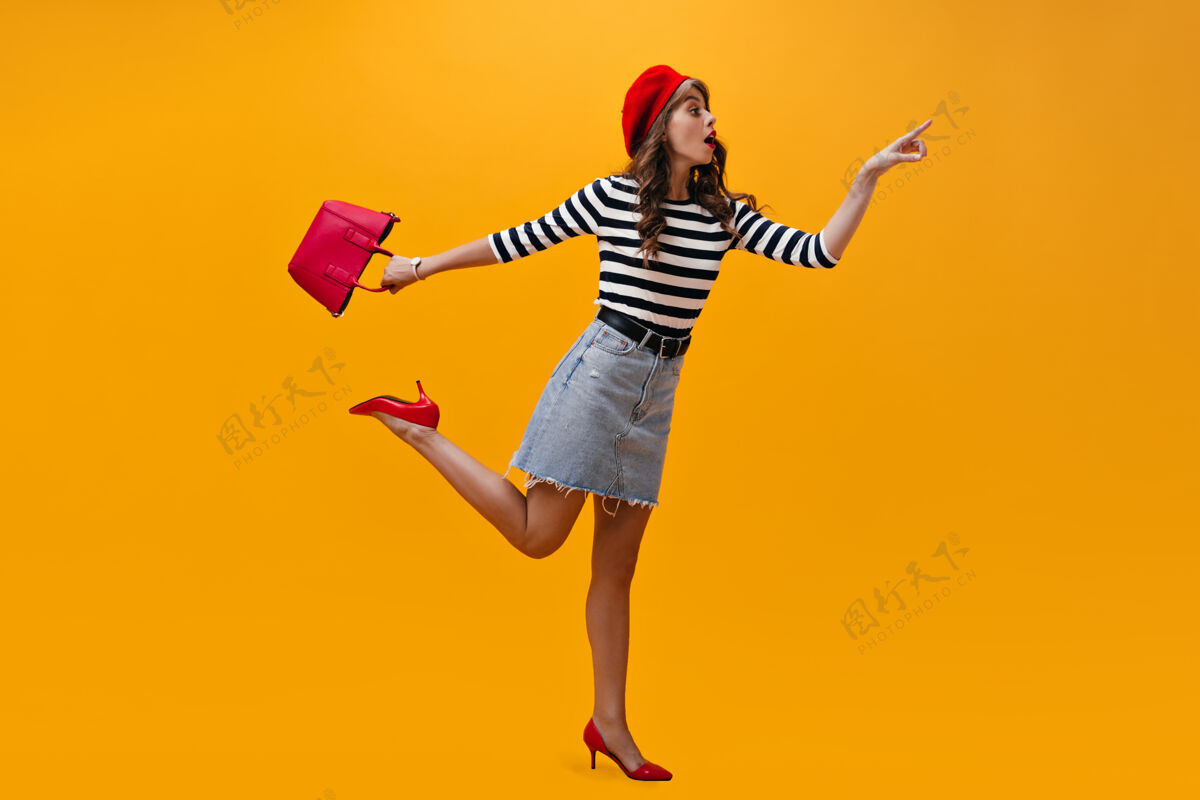 成人穿着条纹衬衫的女人在橙色背景上摆出令人惊讶的姿势穿着红色贝雷帽和时髦裙子的酷女孩很有趣橙色微笑空间