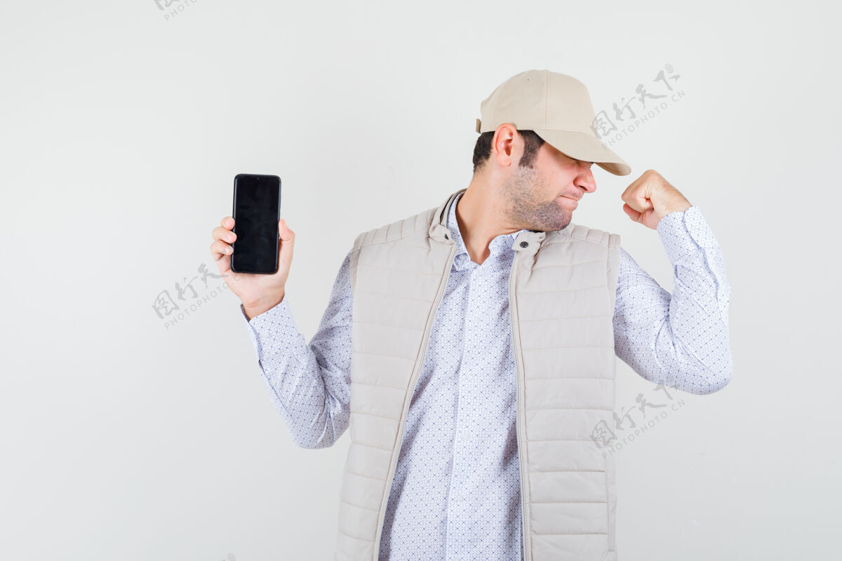 帽子年轻人一只手拿着电话 摆出优胜者的姿势 穿着米色夹克 戴着帽子 看上去很乐观正面图成人显示男子