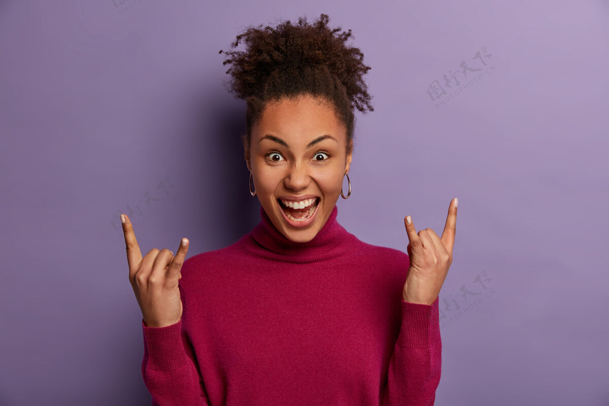 微笑兴奋欢快的卷发女摇滚歌手做重金属标志 在很棒的摇滚音乐会上玩得很开心 感觉很酷 穿着休闲的马球衫 看起来很自信 在紫色的墙壁上摆出室内姿势摇滚音乐会卷发