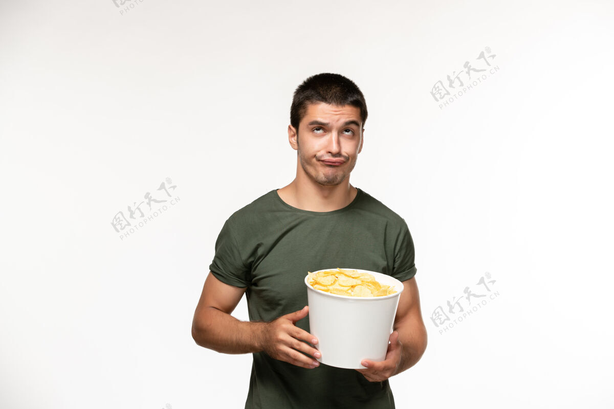 年轻男性正面图身穿绿色t恤的年轻男性手持土豆cips在浅白的墙上看电影人孤独的电影杯子土豆观看