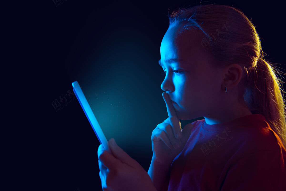 情绪严肃白种人女孩的肖像在霓虹灯下被隔离在黑暗的墙上漂亮的女模特使用平板电脑人类情感的概念 面部表情 销售 广告 现代科技 小玩意成功手势员工