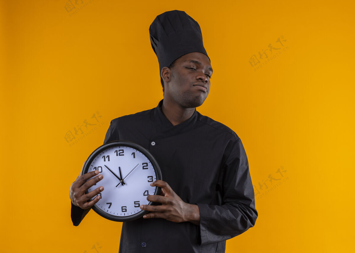 复制穿着厨师制服的年轻恼怒的美国黑人厨师拿着时钟 看着橙色的一边 留着复印空间壁板橙色时钟