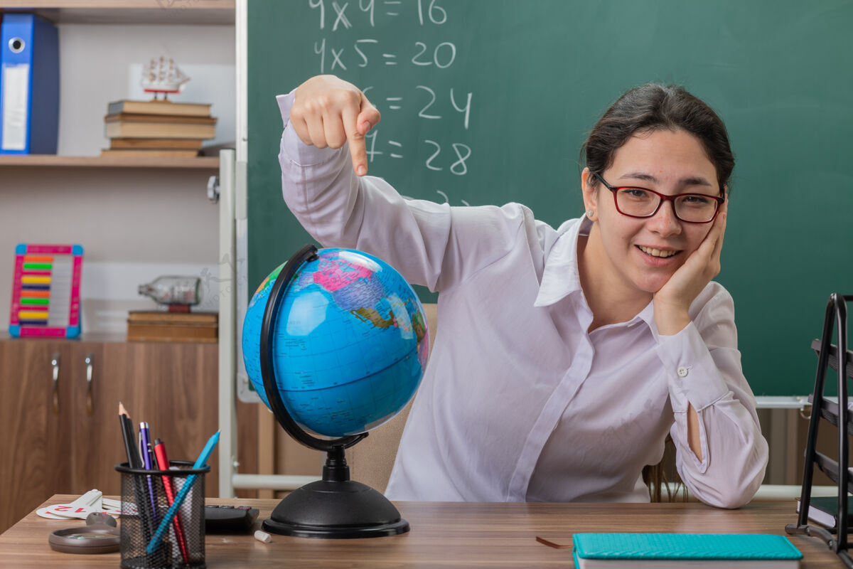 拿着年轻的女老师戴着眼镜拿着地球仪用食指指着地球仪兴高采烈地笑着解释课坐在教室黑板前的课桌上欢呼地球仪微笑