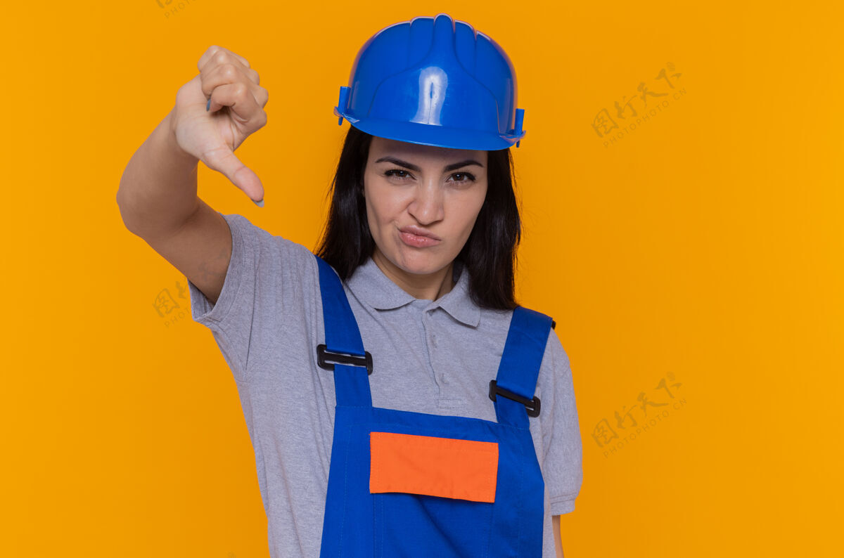 制服身穿建筑制服 头戴安全帽的年轻建筑工人不高兴地朝前看 拇指朝下 站在橙色的墙上展示倒下建筑工人