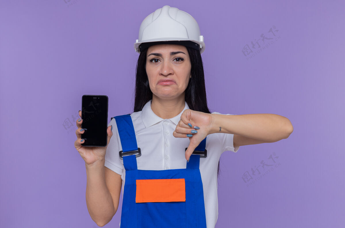 向下身穿建筑制服 头戴安全帽的年轻建筑女工人展示着智能手机 不高兴地看着前面 大拇指朝下站在紫色的墙上不高兴站着建筑