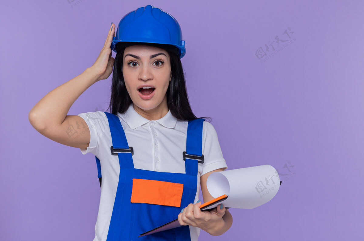 铅笔身穿建筑制服 戴着安全帽 拿着写字板和铅笔的年轻建筑妇女站在紫色的墙上 惊讶地看着前面剪贴板制服建筑