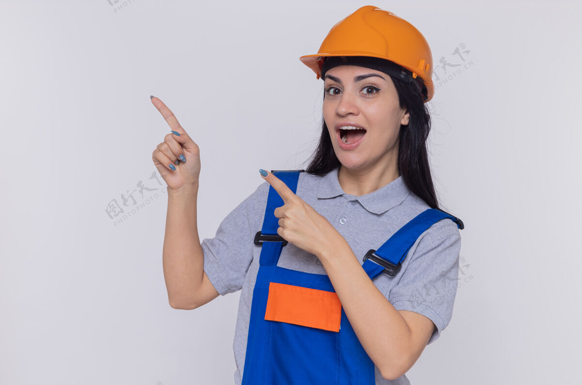 头盔身穿施工制服 头戴安全帽的年轻建筑工人望着前方 开心而积极地微笑着 用食指指着站在白墙上的一侧指向站安全