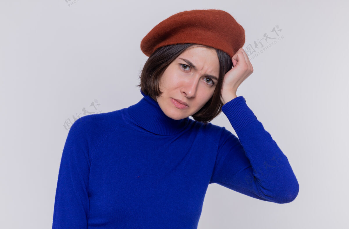 手穿着蓝色高领毛衣 头戴贝雷帽 头发短发的年轻女子站在白色的墙壁上 困惑地看着前面 非常焦急地把手放在头上贝雷帽头女人