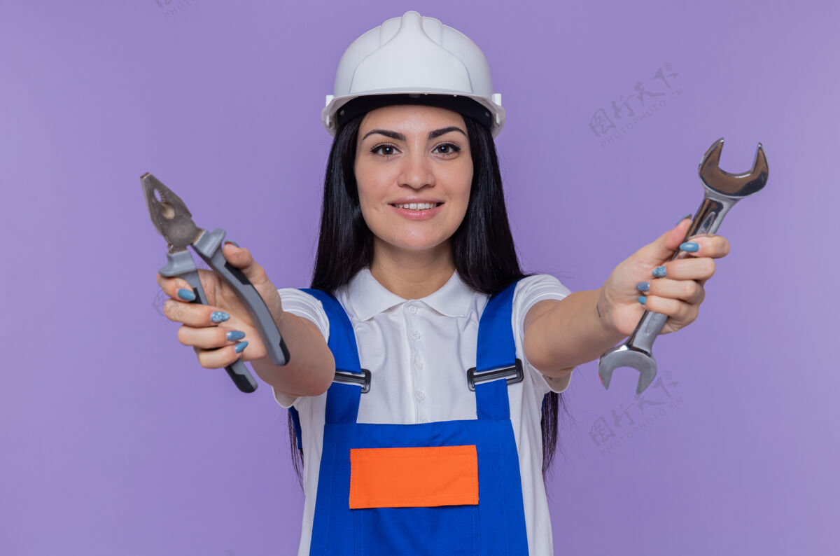 制服身穿建筑制服 头戴安全帽 手持扳手和钳子的年轻建筑妇女站在紫色的墙上 微笑着自信地看着前方女人自信头盔