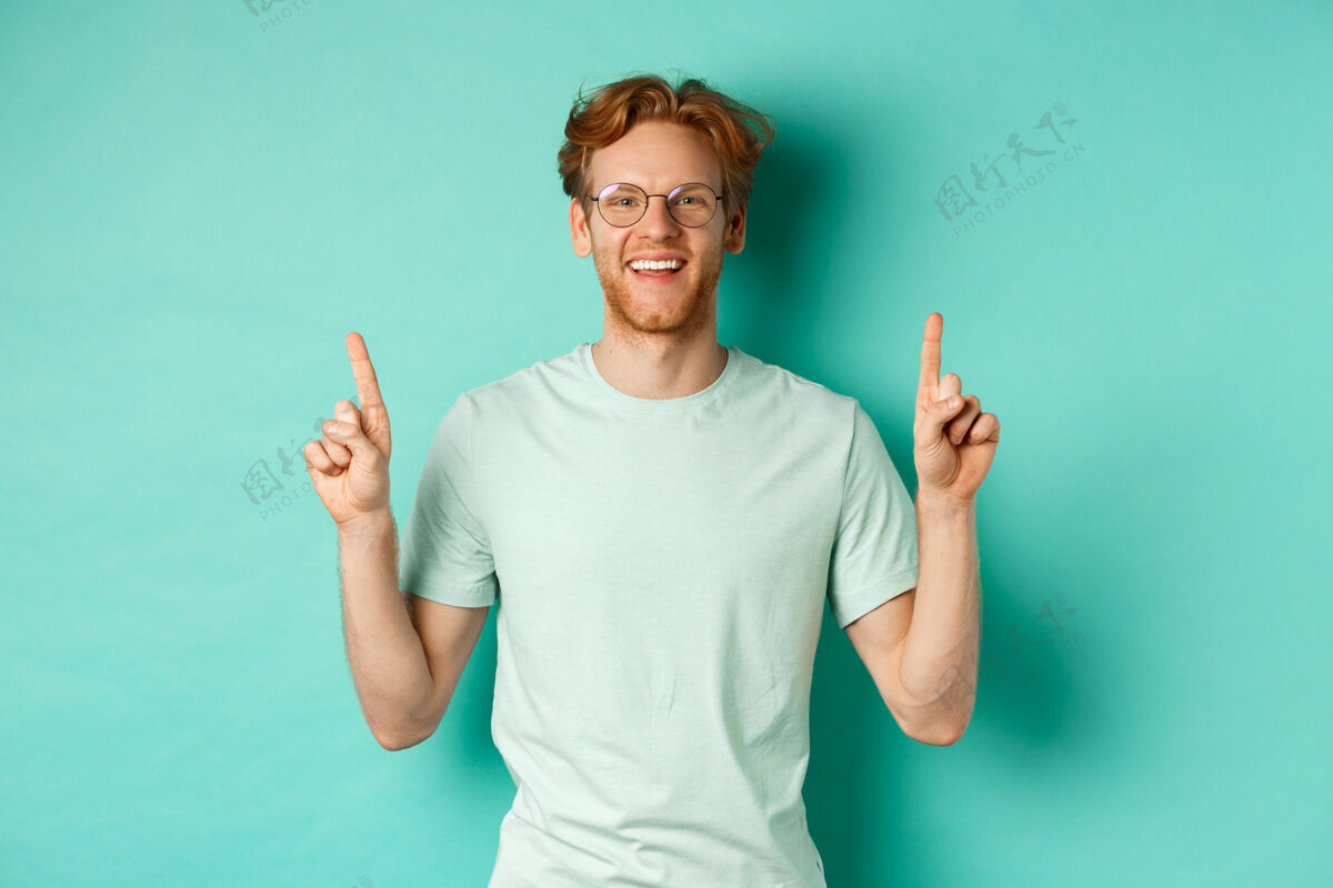 男性一个留着红头发 戴着t恤和眼镜的帅哥 微笑着 手指朝上 展示促销品 站在绿松石背景上头发成人T恤