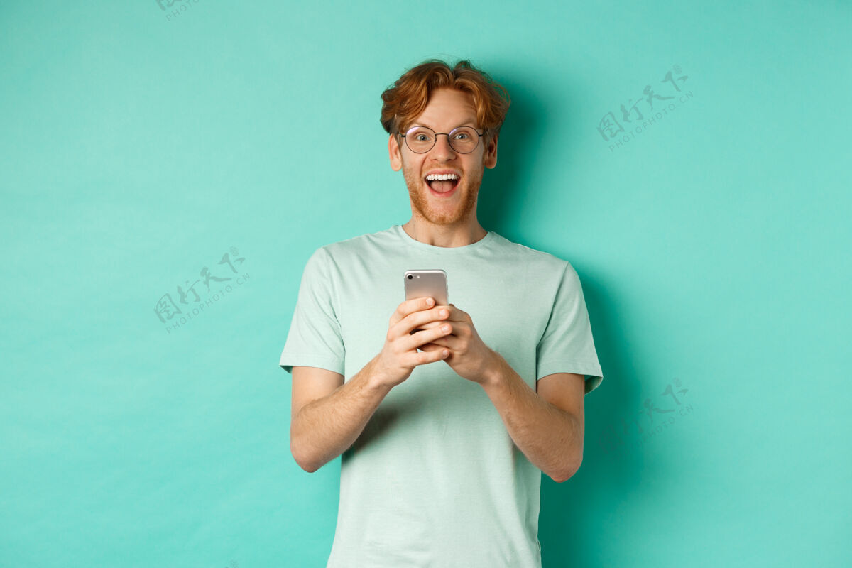 沟通一个戴着眼镜的红发男人站在绿松石色的背景下 看了智能手机上的广告后 惊讶地看着相机男信息微笑