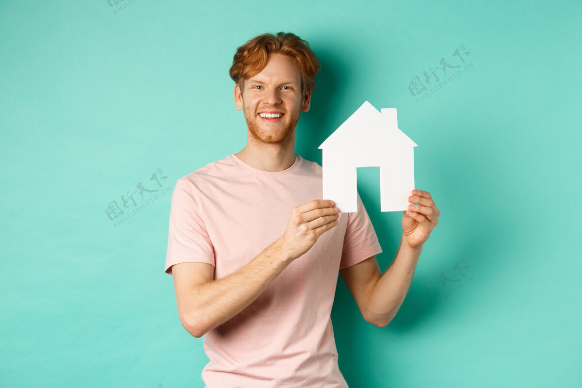 姜房地产概念年轻人红发 穿着t恤 展示纸房子剪贴画 微笑着快乐 站在薄荷背景下男人学生财产