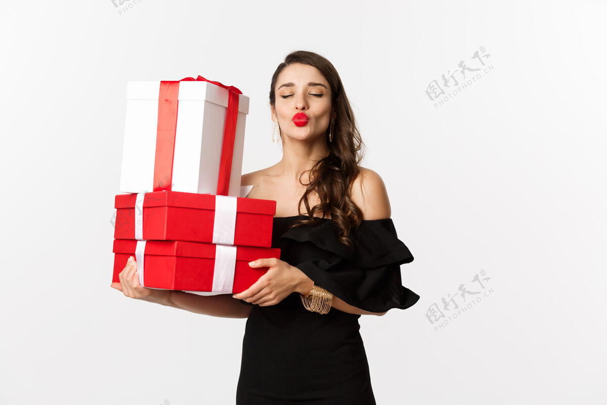 亲吻庆祝和圣诞节的概念愚蠢的女人穿着优雅的黑色连衣裙 拿着圣诞节和新年礼物 撅着嘴唇亲吻 快乐地站在白色背景上优雅女性美容