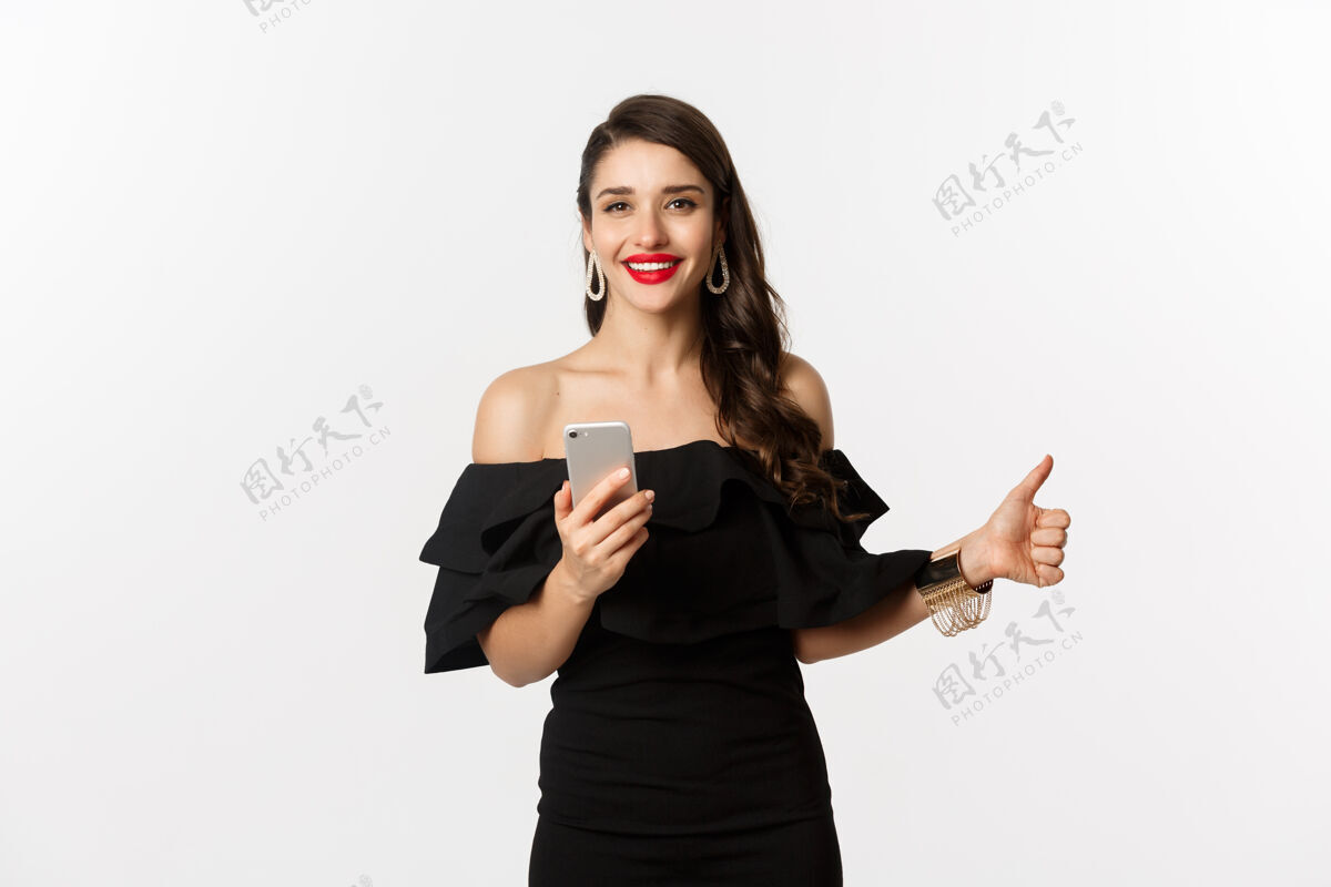 年轻网上购物概念穿着时髦的黑色连衣裙 化妆 竖起大拇指 使用手机应用程序 白色背景的女人女性Brunette女人
