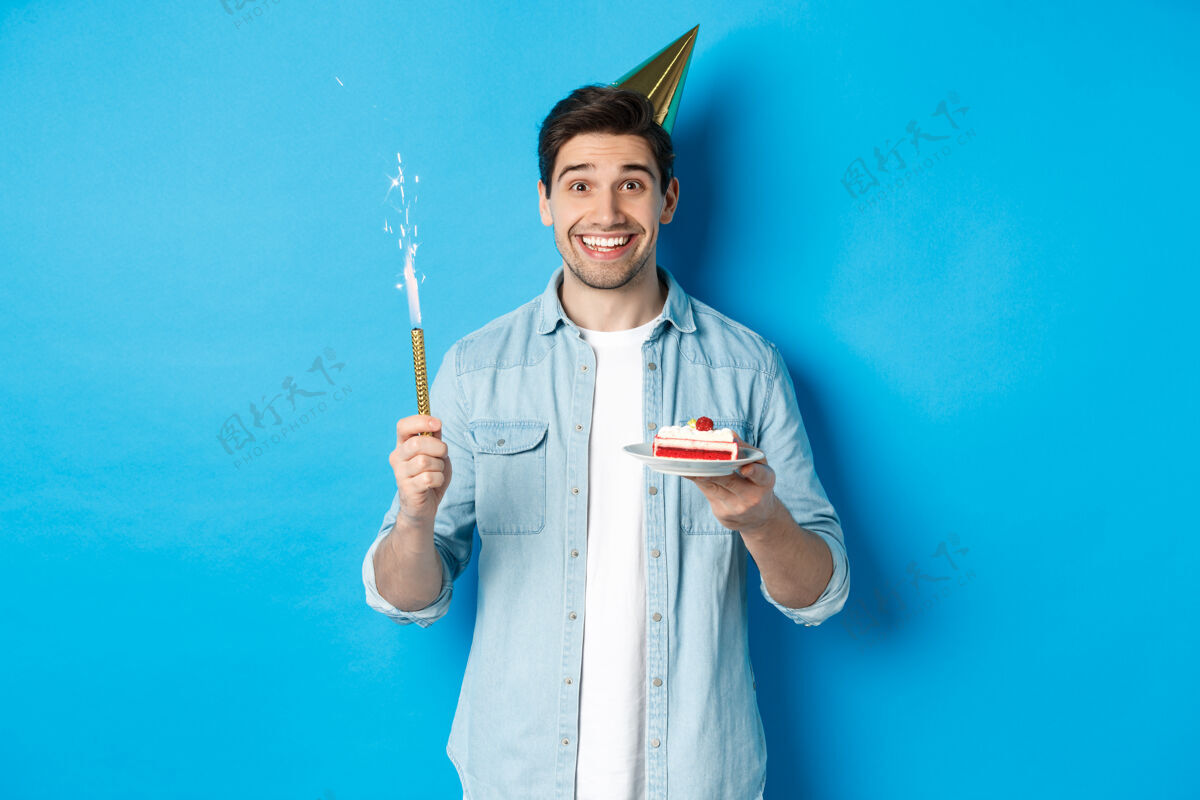休闲快乐的年轻人戴着派对帽庆祝生日 手里拿着生日蛋糕 微笑着站在蓝色的背景上时尚生日模特