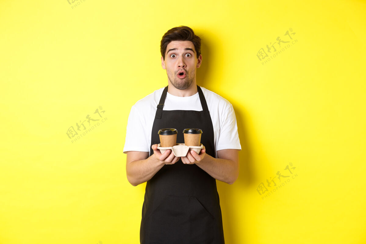 黄色咖啡师端着咖啡 惊讶地看着镜头 穿着黑色围裙 站在黄色背景下年轻工作室帅哥