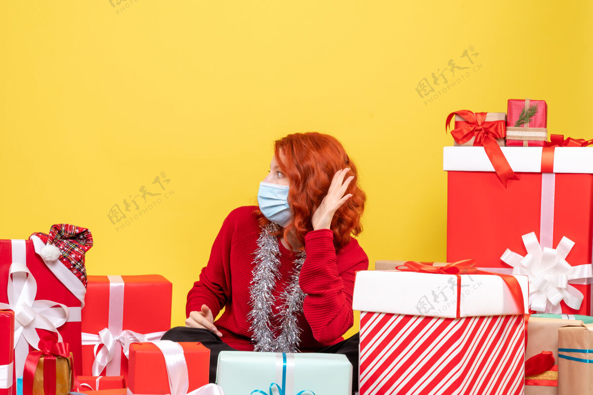 前面正面图：坐在圣诞礼物旁边戴着黄色面具的年轻女子礼物外科口罩病毒